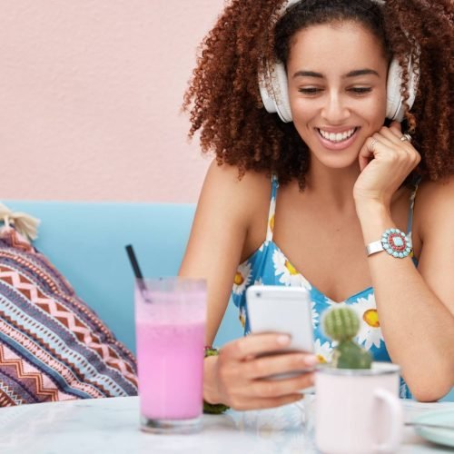 Horizontal shot of pleased African American dark skinned woman listens music from radio website, connected to wireless internet, drink smoothie and eats delicious cake, sits on sofa in coffee shop Horizontal shot of pleased African American dark skinned woman listens music from radio website, connected to wireless internet, drink smoothie and eats delicious cake, sits on sofa in coffee shop